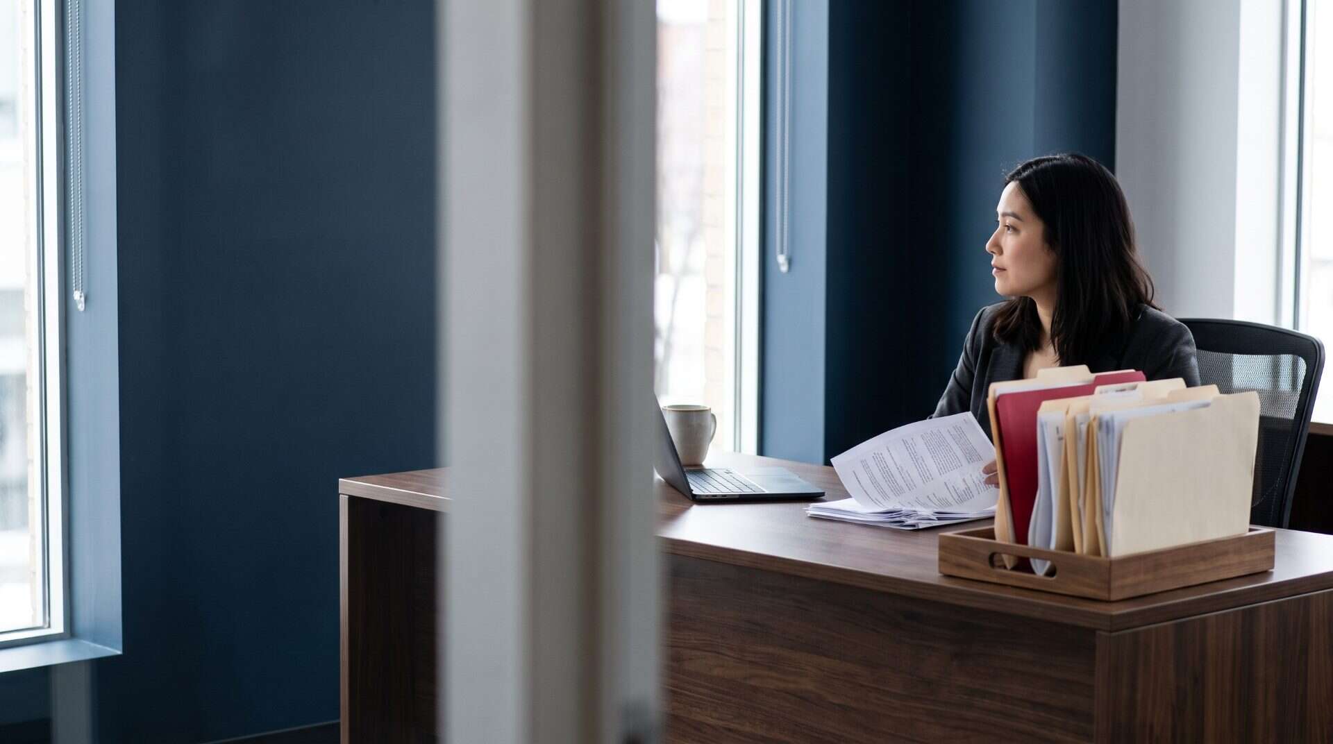 Operations analyst at a walnut desk holding a printed invoice, a tray of sorted folders with one crimson folder beside her, navy office walls behind