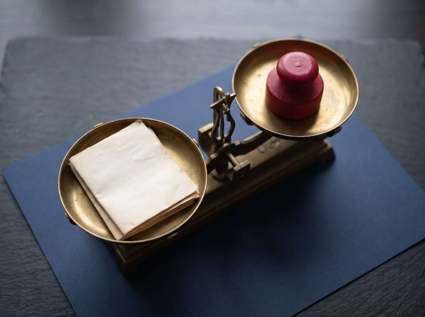 Brass balance scale on navy paper, a stack of documents in one pan and a crimson weight in the other